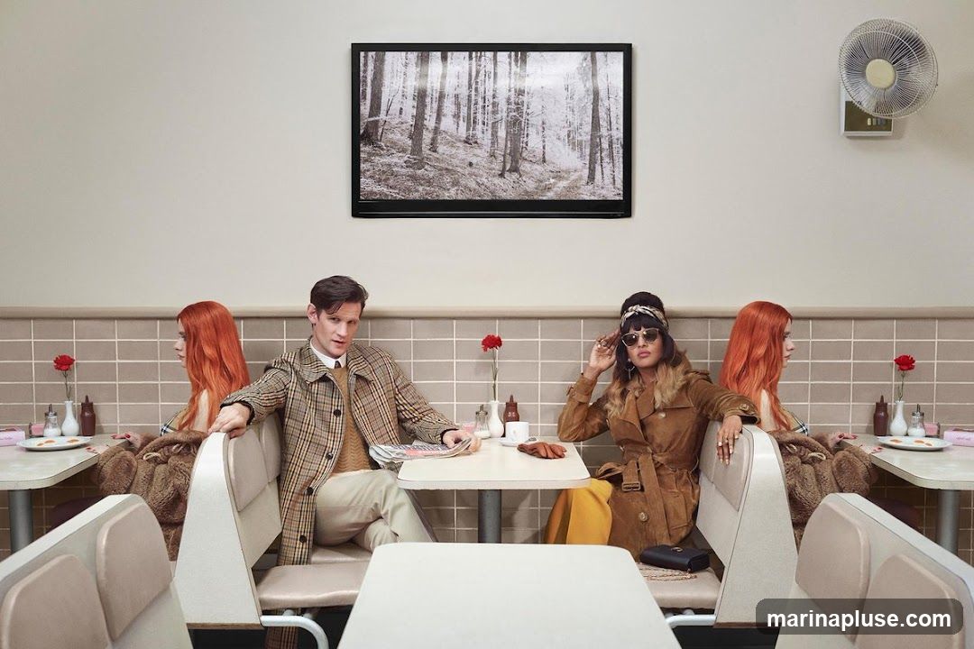 A man and a woman sit in a diner booth, facing away from each other. The man wears a plaid coat and a sweater, while the woman wears a brown suede coat and sunglasses. A framed picture of a snowy forest hangs on the wall above them. The diner has tiled walls and booth seating. There are condiments and a small plate with food on the tables.