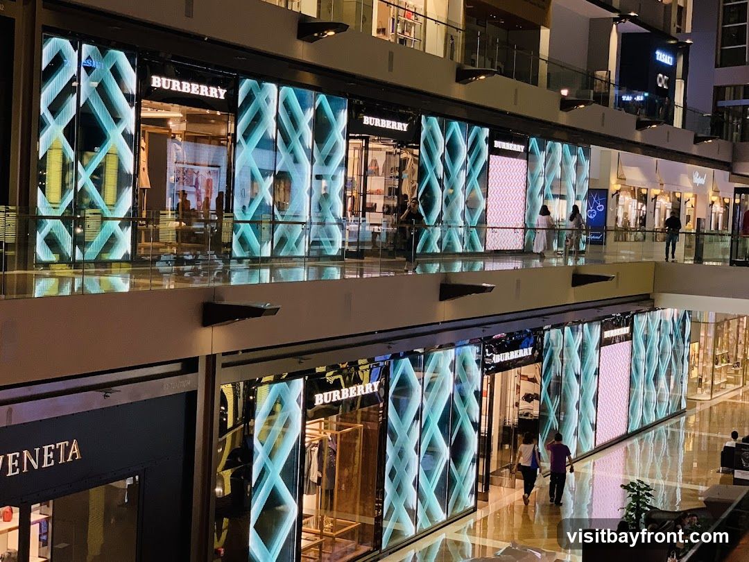 A view of multiple Burberry storefronts within a modern shopping mall. The stores feature large, illuminated displays with geometric patterns. People are seen walking on the upper and lower levels of the mall, with some browsing near the shops. Other luxury brands are also visible in the background.