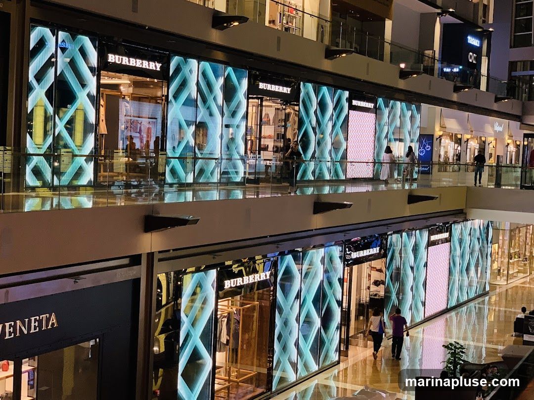 A view of multiple Burberry storefronts within a modern shopping mall. The stores feature large, illuminated displays with geometric patterns. People are seen walking on the upper and lower levels of the mall, with some browsing near the shops. Other luxury brands are also visible in the background.