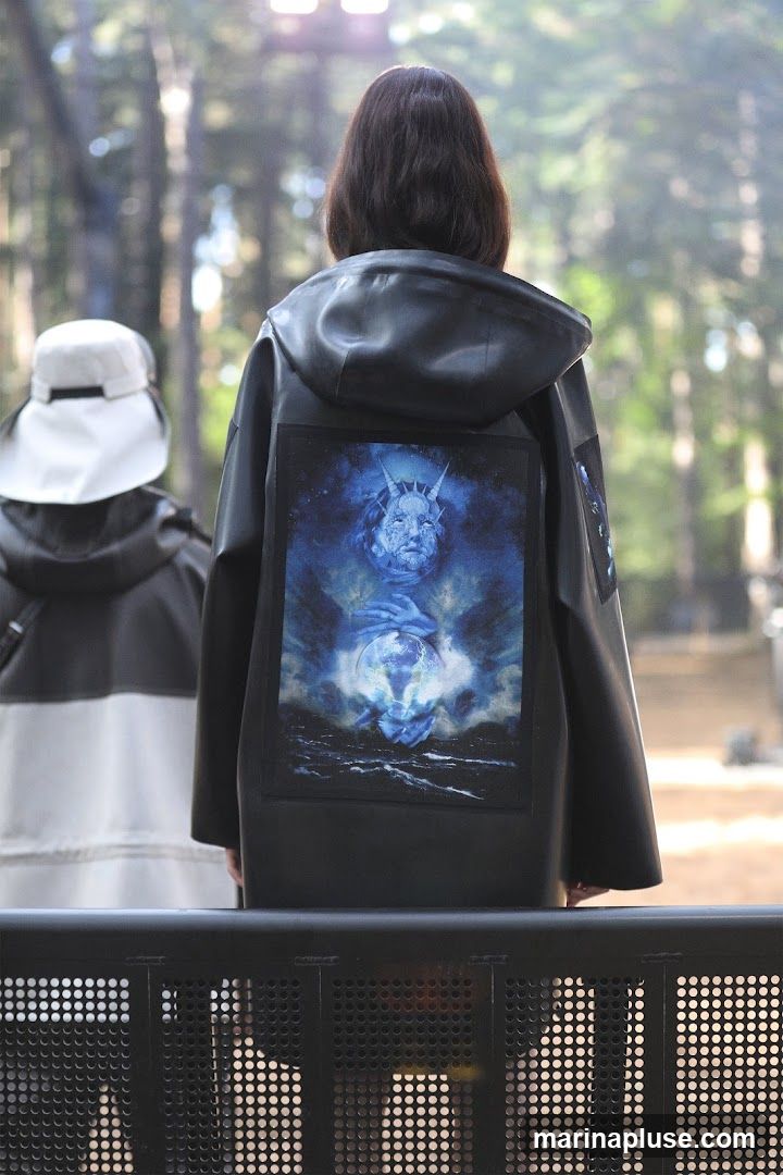 The back of a person wearing a dark jacket with a large, detailed graphic on it. The graphic depicts a blue-toned image of a face with horns holding a globe. Another person wearing a white hat is visible to the left. The background is blurred and appears to be a forest or outdoor setting.