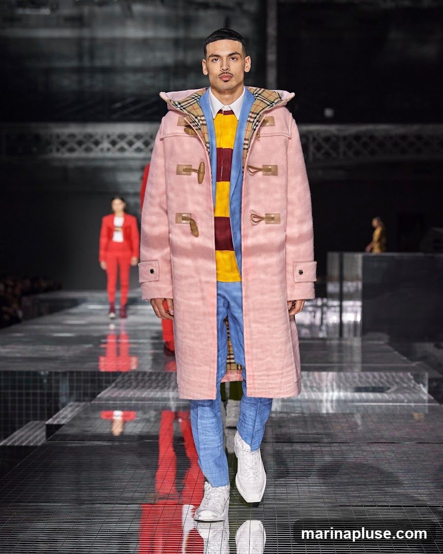 A male model walks down a runway during a fashion show. He is wearing a pink duffle coat with a plaid lining, a yellow and burgundy striped shirt, blue pants, and white sneakers. Other models in red outfits are visible in the background. The runway has a reflective surface.
