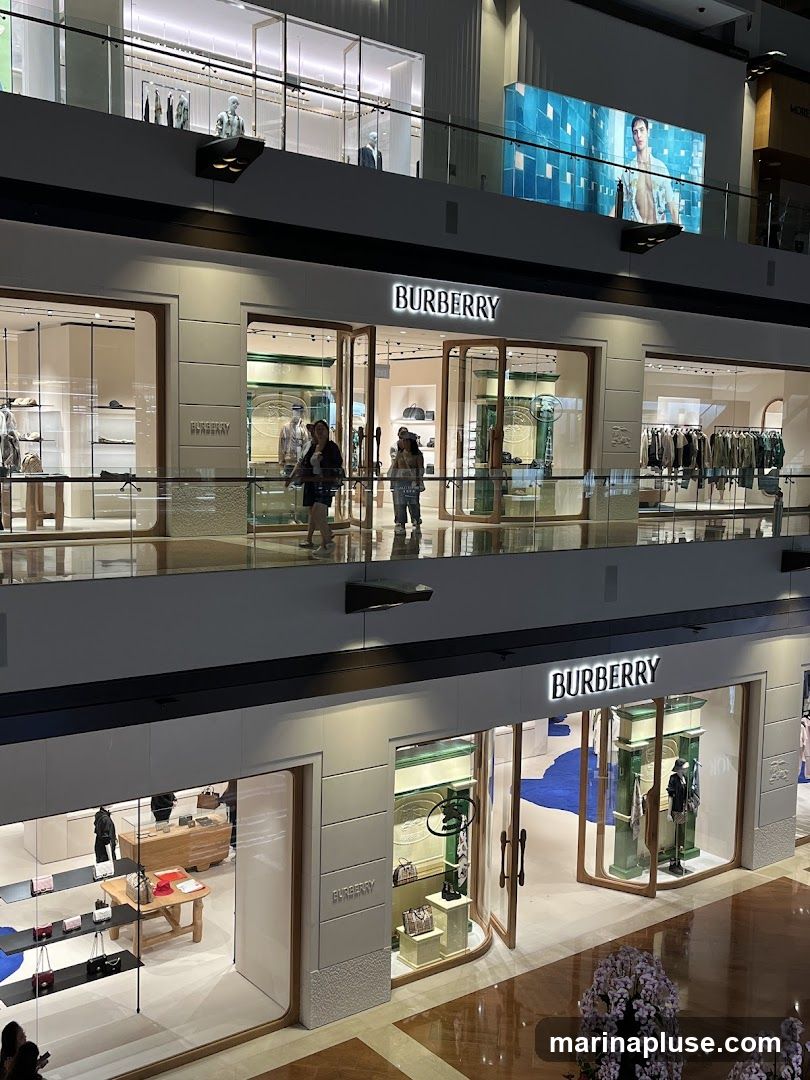 A multi-level interior view of a Burberry store within a shopping mall. The storefronts are visible on two levels, with prominent illuminated "BURBERRY" signage. The upper level shows mannequins displaying clothing behind glass windows. The lower level features displays of handbags and clothing, with customers browsing inside. The overall aesthetic is modern and luxurious, with clean lines and well-lit displays.
