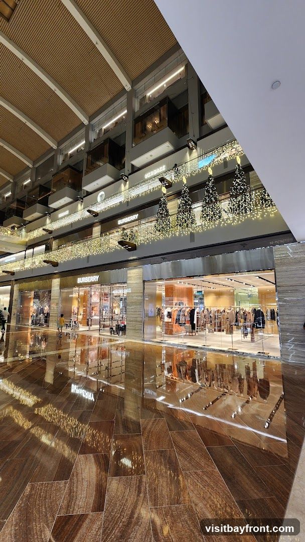 A wide shot of a Burberry store entrance in a modern shopping mall. The store has a glass facade with mannequins displaying clothing inside. The Burberry logo is visible above the entrance. The floor is highly reflective, showing the lights and decorations of the mall, including Christmas trees adorned with lights on the upper levels. Other store signs, such as Versace, are visible on the upper floors.