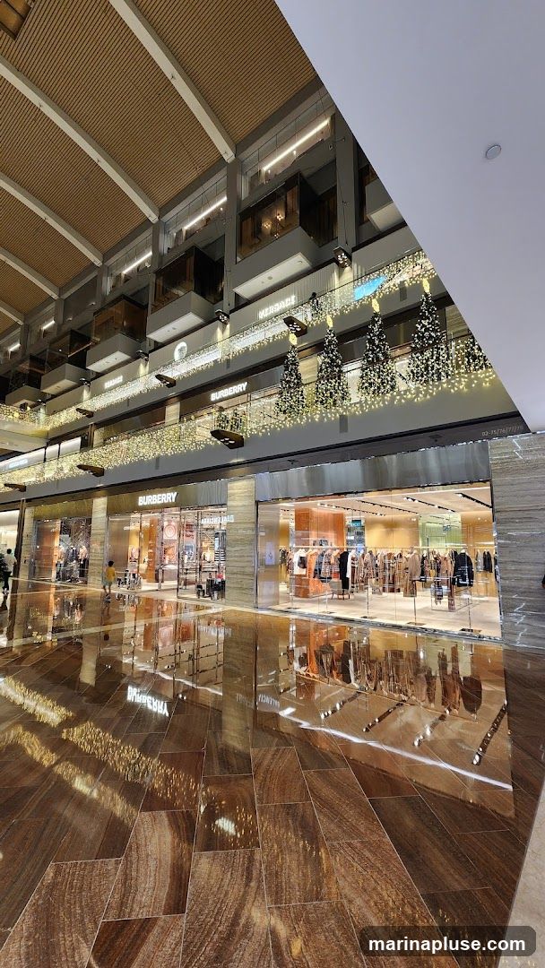 A wide shot of a Burberry store entrance in a modern shopping mall. The store has a glass facade with mannequins displaying clothing inside. The Burberry logo is visible above the entrance. The floor is highly reflective, showing the lights and decorations of the mall, including Christmas trees adorned with lights on the upper levels. Other store signs, such as Versace, are visible on the upper floors.