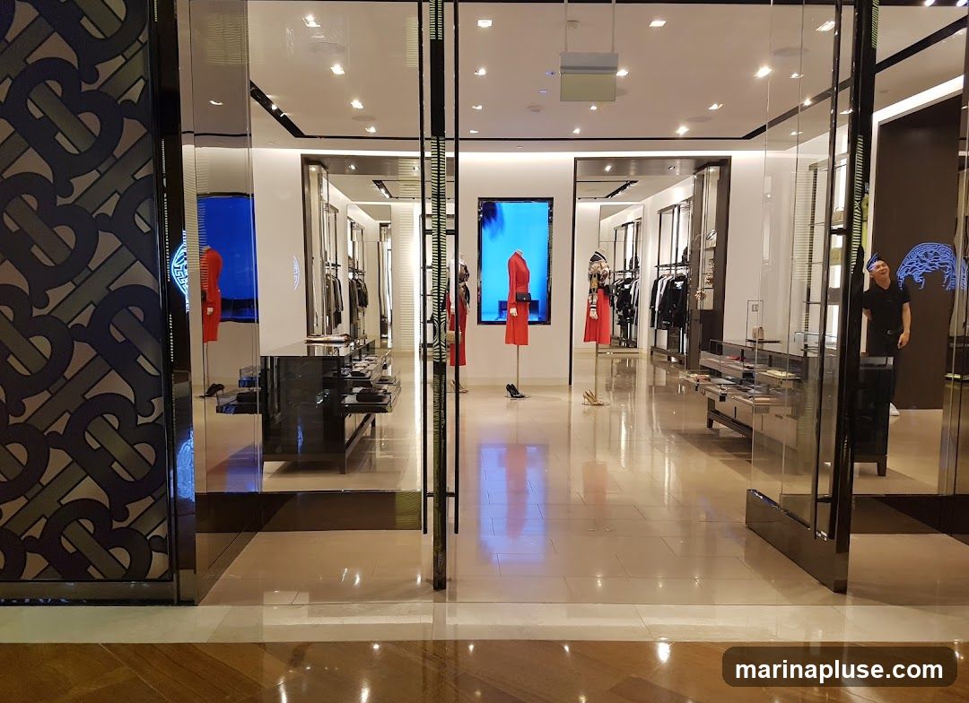The image shows the interior of a high-end fashion store. Racks of clothing, including dresses and jackets, are displayed. Mannequins are dressed in stylish outfits. Digital screens showcase fashion items. The store features a modern and sleek design with polished floors and ample lighting. A person is visible in the reflection of a glass panel.