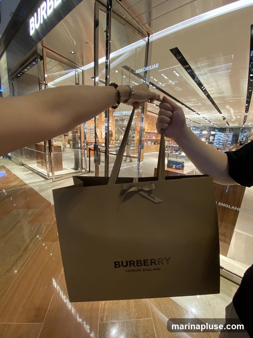 A shopping bag with the Burberry logo is held in front of a Burberry store. The store has a modern glass facade with the Burberry logo visible above the entrance. Inside, merchandise is displayed on shelves.