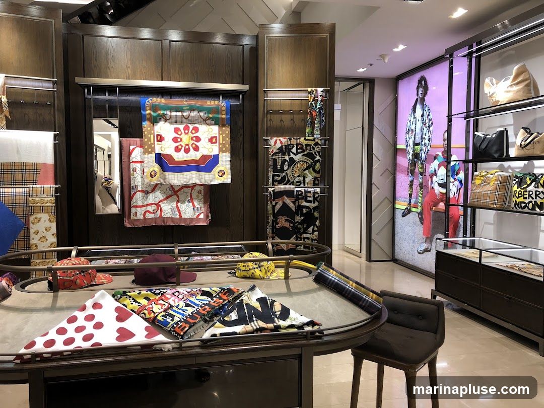 A well-lit retail store interior showcasing a collection of luxury scarves and handbags. Scarves with various patterns, including plaid, floral, and map prints, are displayed on racks and a central table. Handbags and tote bags, some featuring plaid patterns and the text "BURBERRY LONDON", are arranged on shelves. A large digital display on the right features models wearing fashionable clothing.
