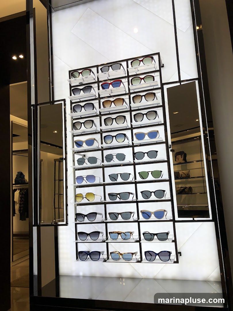 A wall display of various sunglasses in a retail store. The sunglasses are arranged on white shelves in a grid pattern. Some sunglasses have dark frames, while others have lighter or colored frames. The display is illuminated from behind, creating a bright and modern aesthetic. To the left, a glimpse of clothing racks and a mirror can be seen, suggesting this is part of a larger fashion boutique.