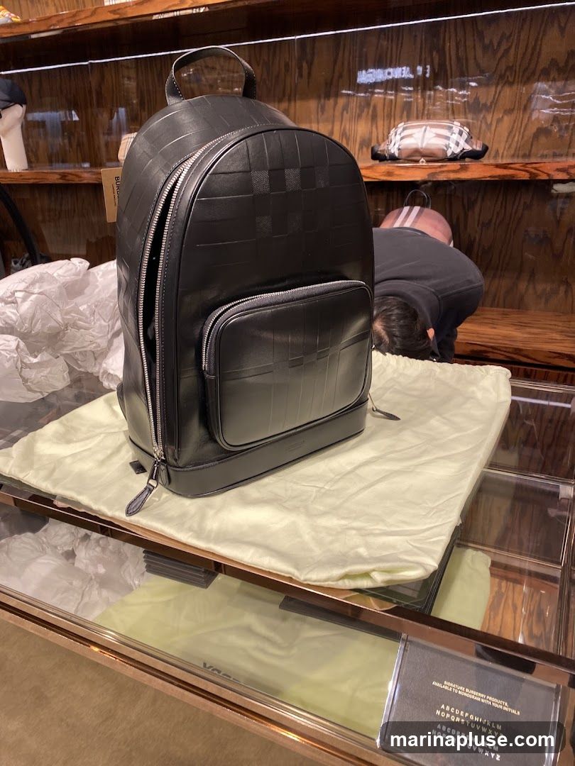 A black leather backpack with a checkered pattern is displayed on a light-colored cloth bag. The backpack has a main zippered compartment and a smaller zippered pocket on the front. In the background, shelves display other items, including a checkered bag and a pink bag. A person is seen bending down in the background. A sign at the bottom right provides information about monogramming Burberry products with initials.