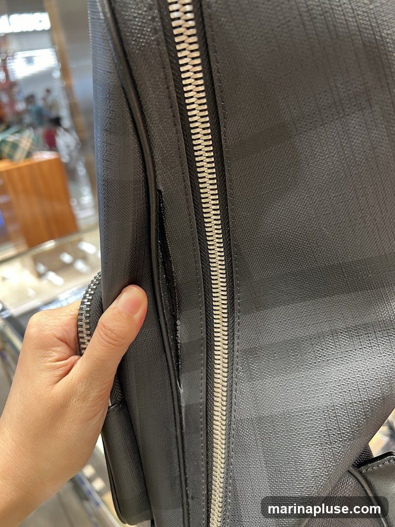 A hand is holding a black bag with a checkered pattern. The bag has a prominent silver zipper. In the blurred background, there are displays and people, suggesting a retail environment.