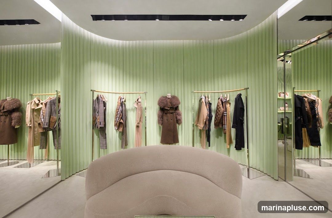 A well-lit, modern luxury fashion store interior with pale green, fluted walls and polished floors. Racks of clothing, including coats, jackets, and dresses, are displayed on gold-toned hangers. Mannequins showcase select garments. A plush, curved sofa is positioned in the foreground. Mirrors line some of the walls, reflecting the merchandise and creating a sense of spaciousness. Shelving units with small items are visible in the background.