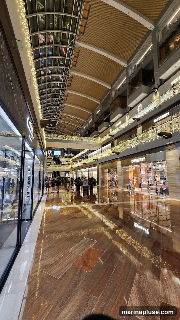 A wide-angle view of an indoor shopping mall walkway. The ceiling is high with a curved, glass-covered section and wooden panels. String lights are draped along the railings of the upper floors, creating a festive atmosphere. The floor is made of polished, multi-colored stone tiles that reflect the lights and the surrounding architecture. Several storefronts are visible on both sides of the walkway, with some featuring large glass windows displaying merchandise. People are walking along the mall. Brands like Burberry and Adidas are visible on storefronts.