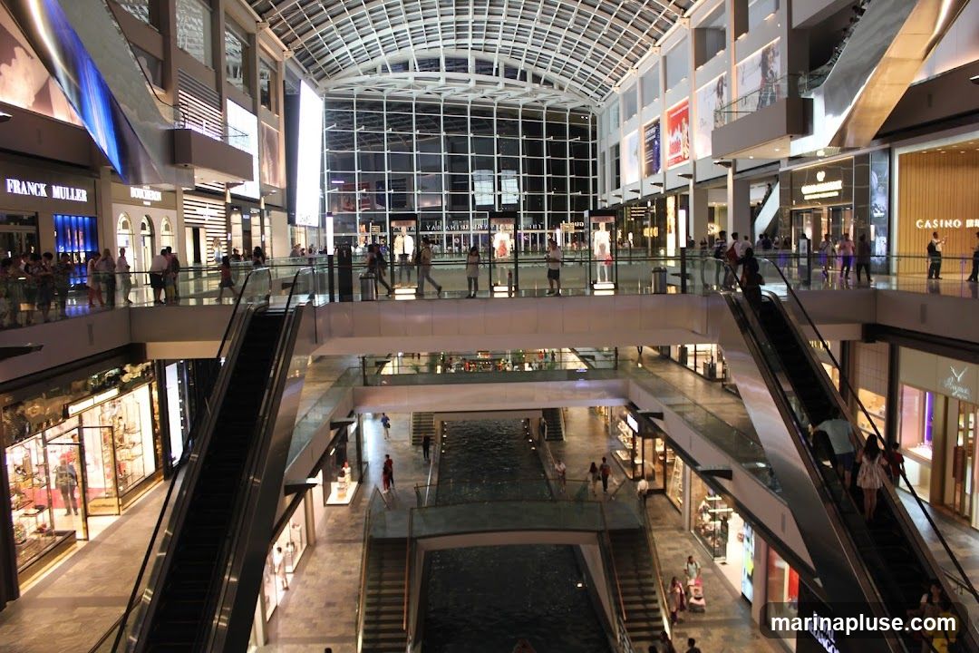 The image shows the interior of a large, modern shopping mall with multiple levels. Escalators and walkways connect different floors, and a canal runs through the center of the lower level. Various shops with illuminated storefronts are visible, including Franck Muller, Boucheron, and a casino. People are seen shopping and walking around. The mall features a glass ceiling that allows natural light to enter.