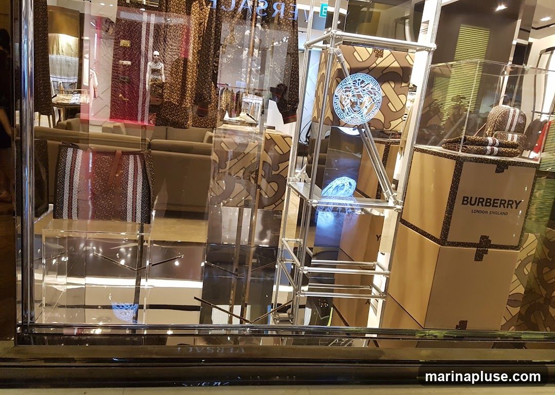 A store window display featuring luxury fashion items. In the foreground, a large tote bag with a patterned design is displayed on a clear stand. To the right, stacked boxes with the Burberry logo and "LONDON ENGLAND" are visible, topped with folded items. A metal structure displays the Versace Medusa logo. The background shows mannequins and furniture, with patterned fabrics and reflections creating a luxurious ambiance.