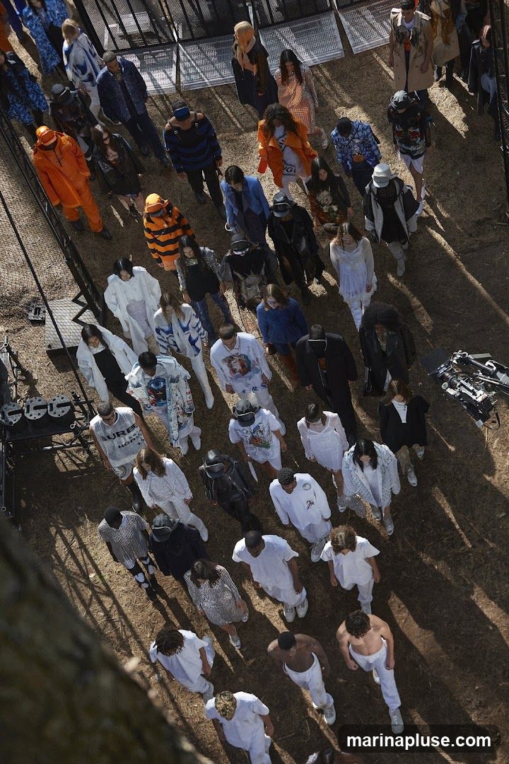 An aerial view of a crowd of people gathered outdoors, possibly for a fashion show or event. The individuals are dressed in a variety of clothing styles and colors, with some wearing bright orange, blue, and striped outfits, while others are in white or dark attire. The ground appears to be dirt or sand, with shadows cast by the people and surrounding structures.