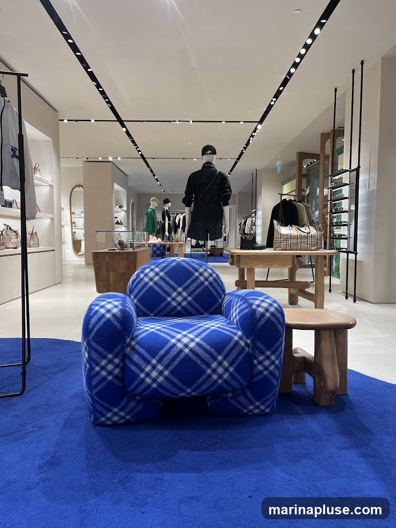 A brightly lit, modern retail space with a focus on fashion. In the foreground, a plush, royal blue armchair with a white plaid pattern sits on a matching blue rug. To the right, a small, organically shaped wooden stool complements a larger wooden display table. The background reveals mannequins dressed in contemporary clothing, including jackets, shorts, and hats. Shelving units display handbags and accessories, with more clothing racks visible further back. The overall ambiance is clean, sophisticated, and designed to showcase high-end merchandise.