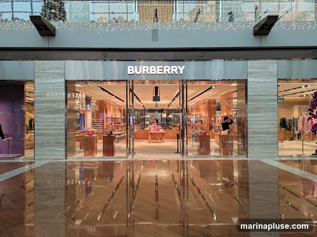 The exterior of a Burberry store with its name prominently displayed above the entrance. The store features large glass windows and doors, revealing a well-lit interior with merchandise displayed on shelves and stands. Reflections of the store and surrounding area are visible on the polished floor.