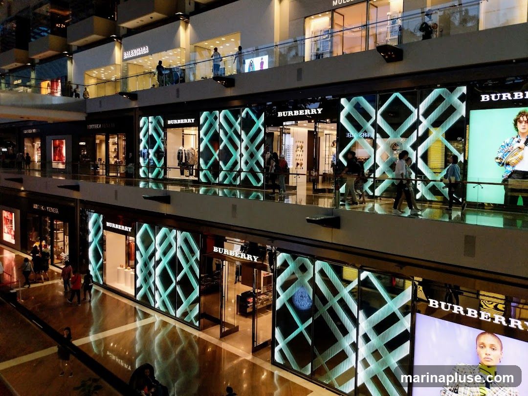 A multi-level indoor shopping mall with several luxury brand stores visible. The image prominently features the Burberry brand with its distinctive plaid pattern displayed on illuminated screens and storefronts. Other brands like Balenciaga and Bottega Veneta are also visible. People are seen walking on the elevated walkways and browsing the shops.