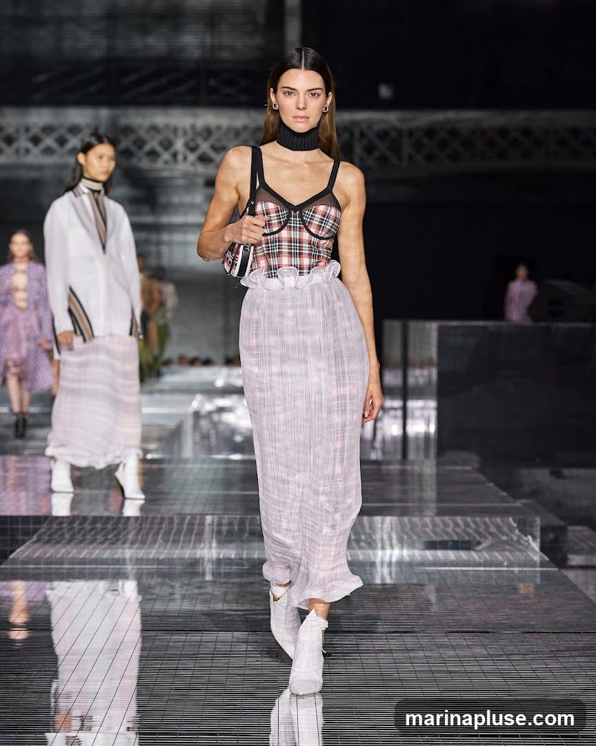 A fashion model walks down a runway during a fashion show. She is wearing a plaid corset top with a ruffled white skirt and white boots. Other models are visible in the background. The runway has a reflective surface.