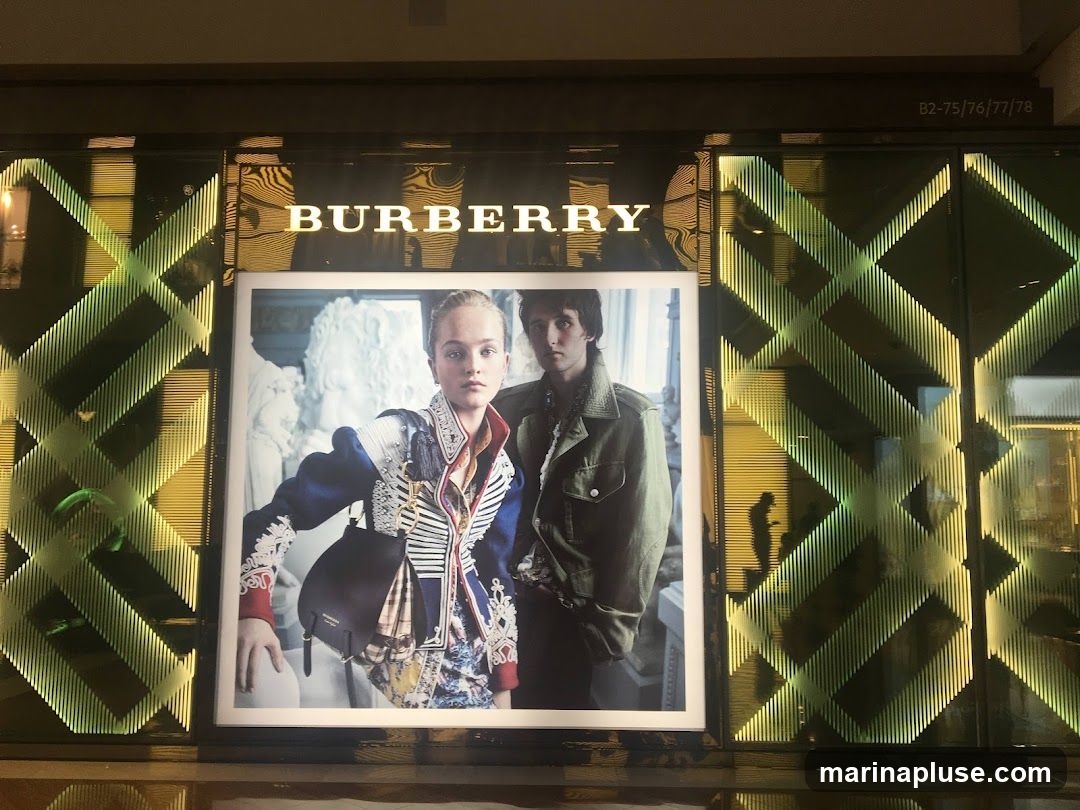 A large advertisement featuring two models is displayed on a store's facade. The Burberry logo is prominently displayed above the advertisement. The storefront features a geometric pattern with green and yellow lighting.