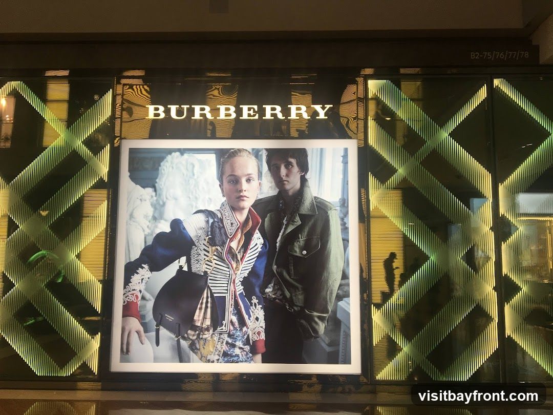 A large advertisement featuring two models is displayed on a store's facade. The Burberry logo is prominently displayed above the advertisement. The storefront features a geometric pattern with green and yellow lighting.