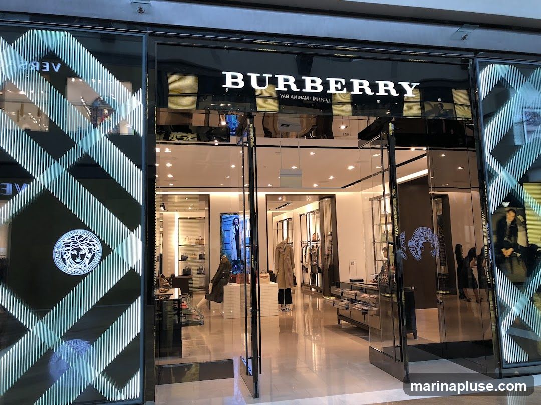 The exterior of a Burberry store with its prominent logo above the entrance. The storefront features large glass windows and doors, with decorative patterns and logos on the glass panels. Inside, clothing and accessories are displayed on shelves and mannequins.