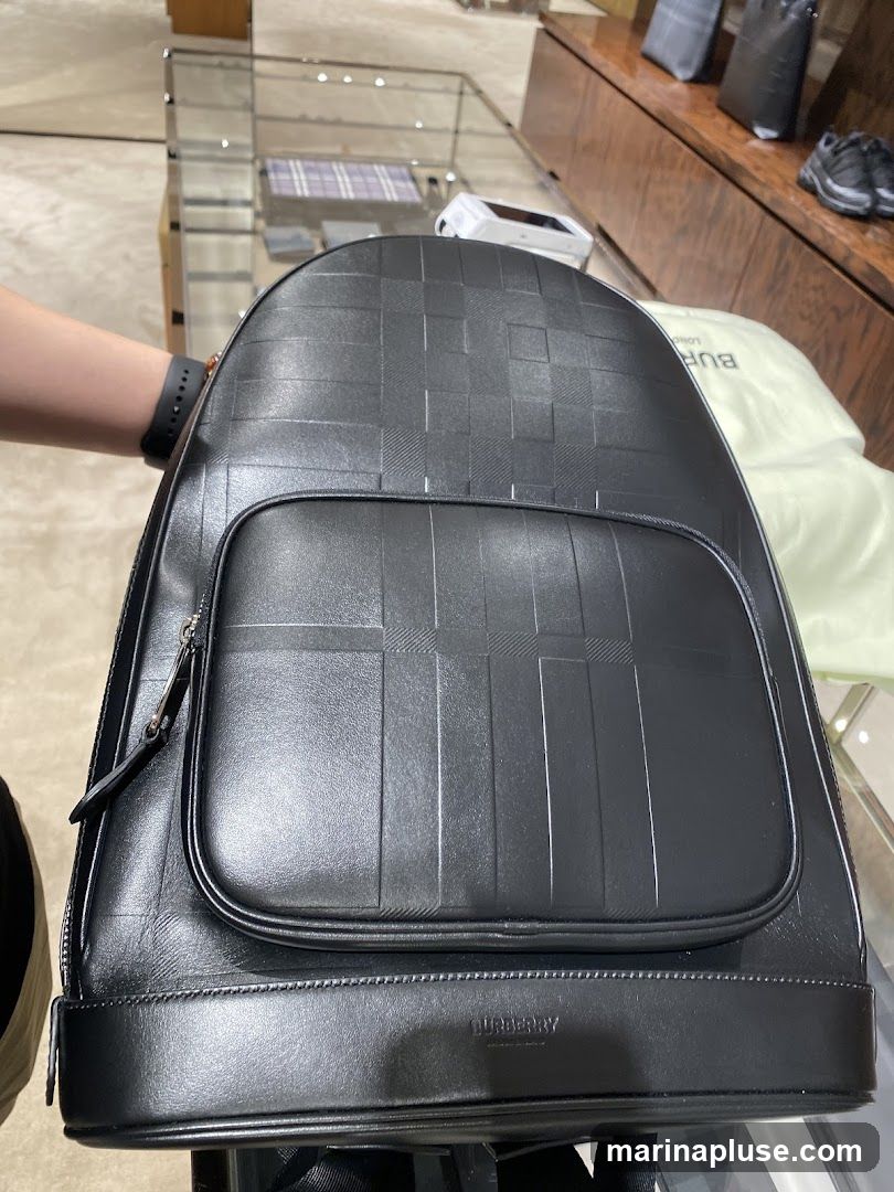 A black leather backpack with a textured pattern is displayed in a store. The backpack has a main compartment and a front pocket with a zipper. The brand name "BURBERRY" is embossed on the bottom front of the backpack. In the background, there are display shelves with other bags and shoes.