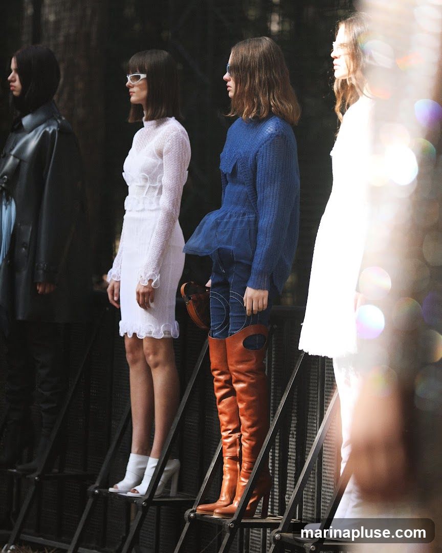 Four models stand in profile on a tiered platform, showcasing outfits. The first model on the left wears a dark coat and pants. The second model is in a white, textured dress with ruffled details and white heels. The third model wears a blue knitted top with a sheer peplum over jeans and tall brown boots. The fourth model is in a white outfit. The background is dark and out of focus, with some bokeh effects on the right side.