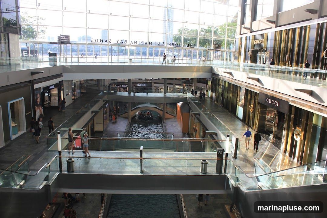 An interior view of a multi-level shopping mall with a canal running through the center. Shops like Chanel and Gucci are visible on the upper levels. People are seen walking on the walkways and a gondola boat is on the canal. The mall has a modern design with glass railings and a large glass ceiling.