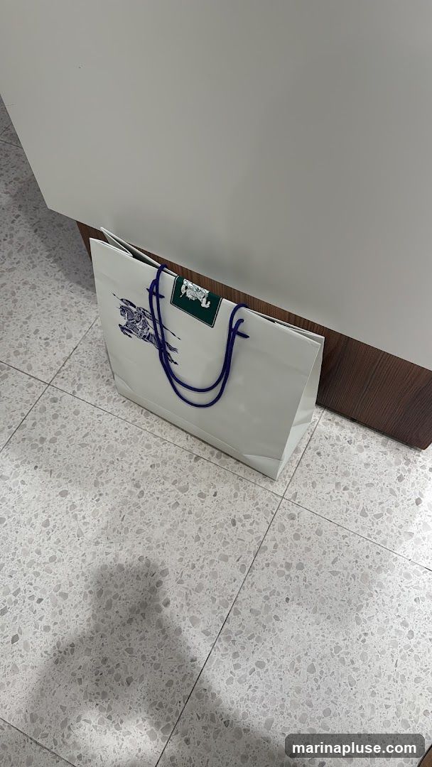 A white shopping bag with blue handles and a dark blue graphic of a horse and rider is placed on a terrazzo floor next to a wooden baseboard. A green rectangular tag with a white logo is attached to the bag's handle.