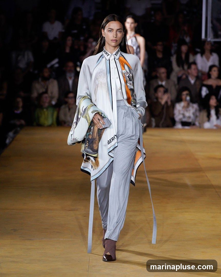 A female model walks down a runway during a fashion show. She is wearing a light blue button-down shirt with a scarf draped over it, featuring animal prints and the word "BURBERRY" repeated along the collar. She also wears light grey trousers and brown heeled shoes. The background shows an audience seated in a dimly lit venue.