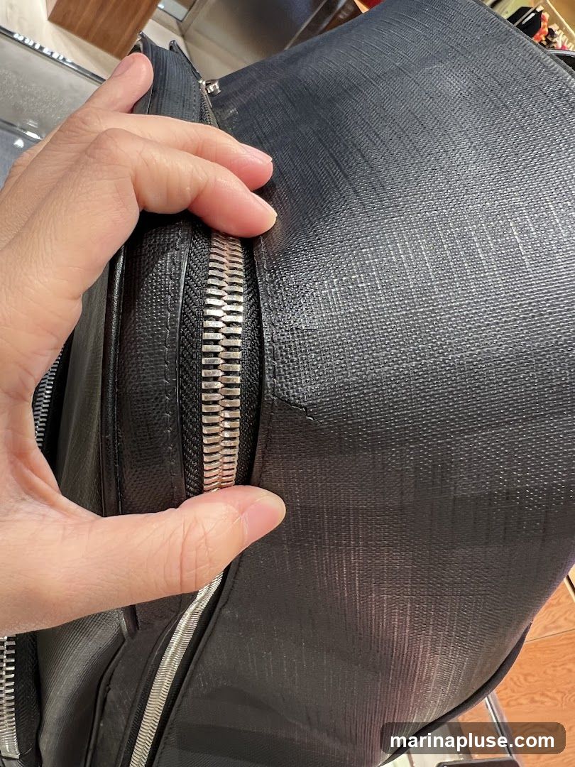 A close-up shot focuses on a person's hand touching the textured surface of a black bag. A prominent silver zipper is visible, partially open. The bag appears to be made of a durable, possibly leather or faux leather material with a subtle crosshatch pattern. The background is blurred, showing hints of shelving and possibly other merchandise, suggesting the item is in a retail environment.