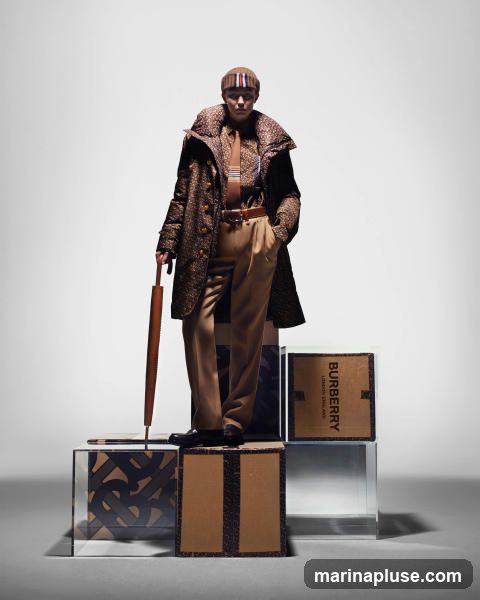 A person stands on stacked boxes, holding an umbrella. They are wearing a patterned Burberry coat, shirt, tie, trousers, and a striped beanie. The boxes are branded with the Burberry logo and pattern.