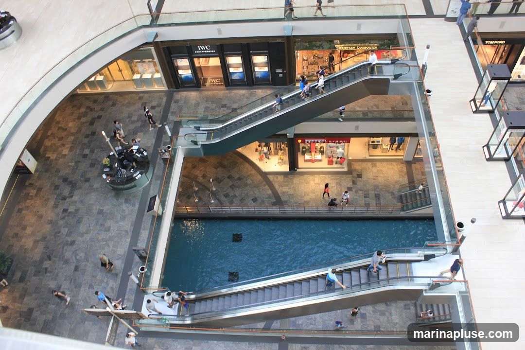 An aerial view of a modern shopping mall interior, featuring multiple levels, escalators, and a central canal filled with blue water. People are seen walking on the floors and using the escalators. Storefronts with visible signage like IWC Schaffhausen and Christian Louboutin are present. Display cases are also visible along the walkways.