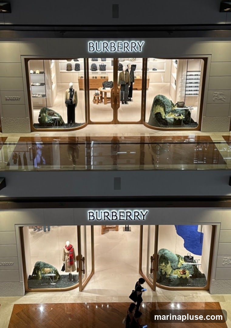 Two levels of a Burberry store are visible, each with a prominent illuminated "BURBERRY" sign above the entrance. The upper level displays mannequins dressed in jackets and trousers, along with handbags and accessories on shelves. The lower level features a mannequin wearing a trench coat and scarf, and a handbag displayed on a table. Both levels have artistic green rock-like installations in the windows.