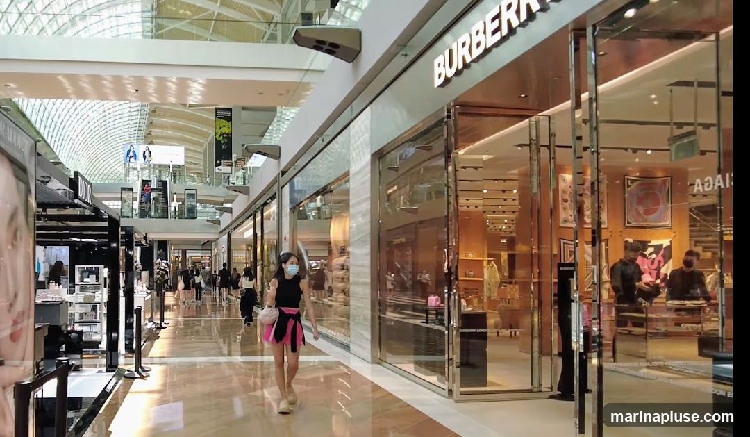 The exterior of a Burberry store in a modern shopping mall. The store has large glass doors and windows, with the Burberry logo prominently displayed above the entrance. Inside, merchandise and displays are visible. People are walking in the mall corridor outside the store.