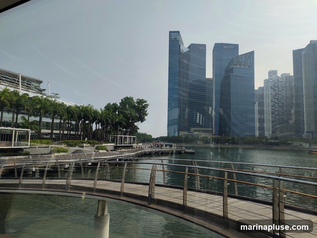 A wide shot of a modern cityscape featuring a curved boardwalk in the foreground, leading towards a waterfront lined with palm trees and contemporary buildings. In the background, several tall skyscrapers dominate the skyline, with visible signage for DBS and HSBC. The water reflects the sky and buildings, creating a serene urban scene.