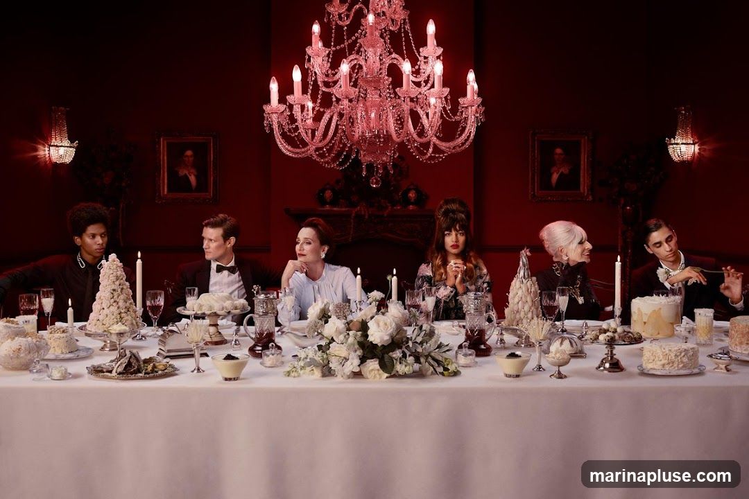 Six people are seated at a long table laden with desserts and floral arrangements. The room is decorated in a dark red hue, with ornate chandeliers and framed portraits on the walls. The individuals are dressed in formal attire, suggesting a special occasion.