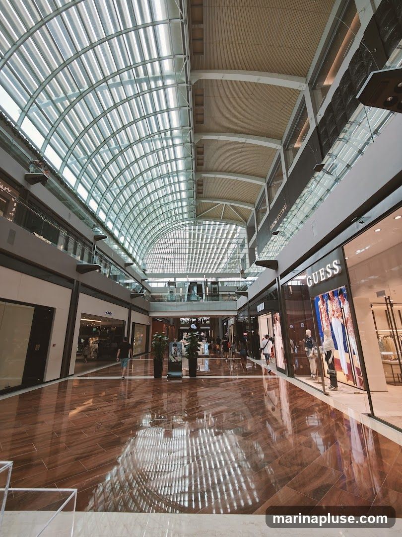 A wide, well-lit corridor inside a modern shopping mall. The ceiling features a large, arched glass roof that allows natural light to filter in, reflecting off the polished wooden floor. On either side of the corridor are various retail stores, including GUESS and ZARA, with their storefronts and displays visible. People are seen walking through the mall, and potted plants add a touch of greenery.