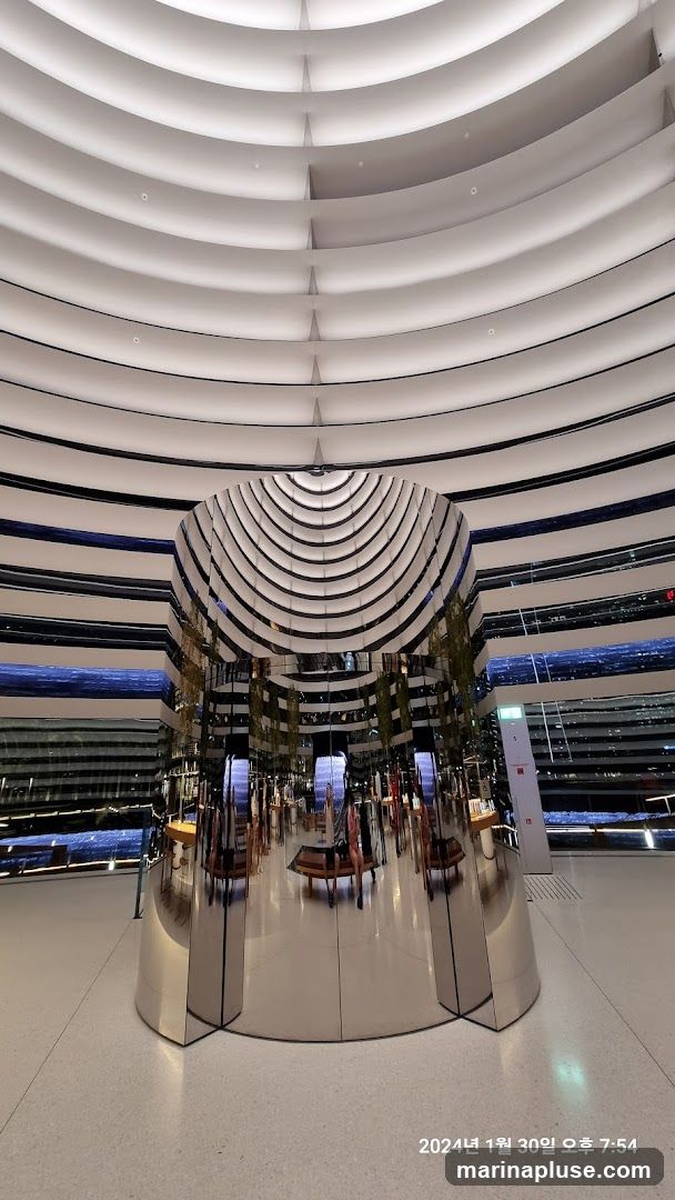 The image shows a spacious, modern interior with a striking, cylindrical, reflective column at its center. The ceiling features a unique design of curved, layered panels with integrated lighting, creating a sense of depth and architectural interest. The reflective column mirrors the surrounding environment, showing glimpses of displays, seating, and people within the space. The walls also have a layered, striped design. The overall impression is one of a contemporary retail or exhibition space.