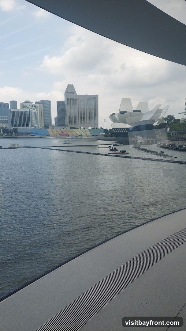 A view of the Singapore skyline across Marina Bay. In the foreground, the water of the bay is visible, with some floating structures and barriers. In the mid-ground, a stadium with colorful seating is visible, along with the distinctive lotus-shaped ArtScience Museum. The background features a cluster of modern skyscrapers, including the Mandarin Oriental hotel. The sky is partly cloudy.