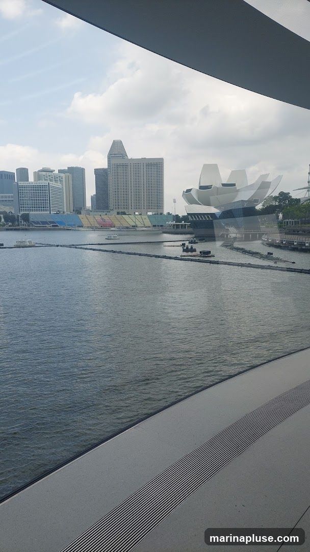 A view of the Singapore skyline across Marina Bay. In the foreground, the water of the bay is visible, with some floating structures and barriers. In the mid-ground, a stadium with colorful seating is visible, along with the distinctive lotus-shaped ArtScience Museum. The background features a cluster of modern skyscrapers, including the Mandarin Oriental hotel. The sky is partly cloudy.