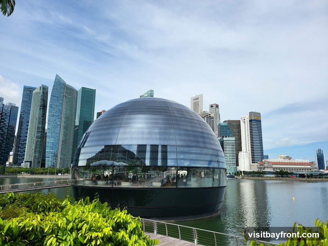 A modern, spherical glass building sits on the edge of a body of water, reflecting the surrounding skyscrapers and a bright blue sky with scattered clouds. Lush green foliage is visible in the foreground, with a walkway and railing. The building's interior appears to be a dining or seating area with people visible inside. The cityscape in the background features numerous tall office buildings.