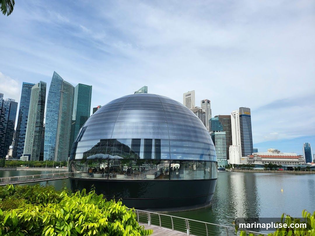 A modern, spherical glass building sits on the edge of a body of water, reflecting the surrounding skyscrapers and a bright blue sky with scattered clouds. Lush green foliage is visible in the foreground, with a walkway and railing. The building's interior appears to be a dining or seating area with people visible inside. The cityscape in the background features numerous tall office buildings.
