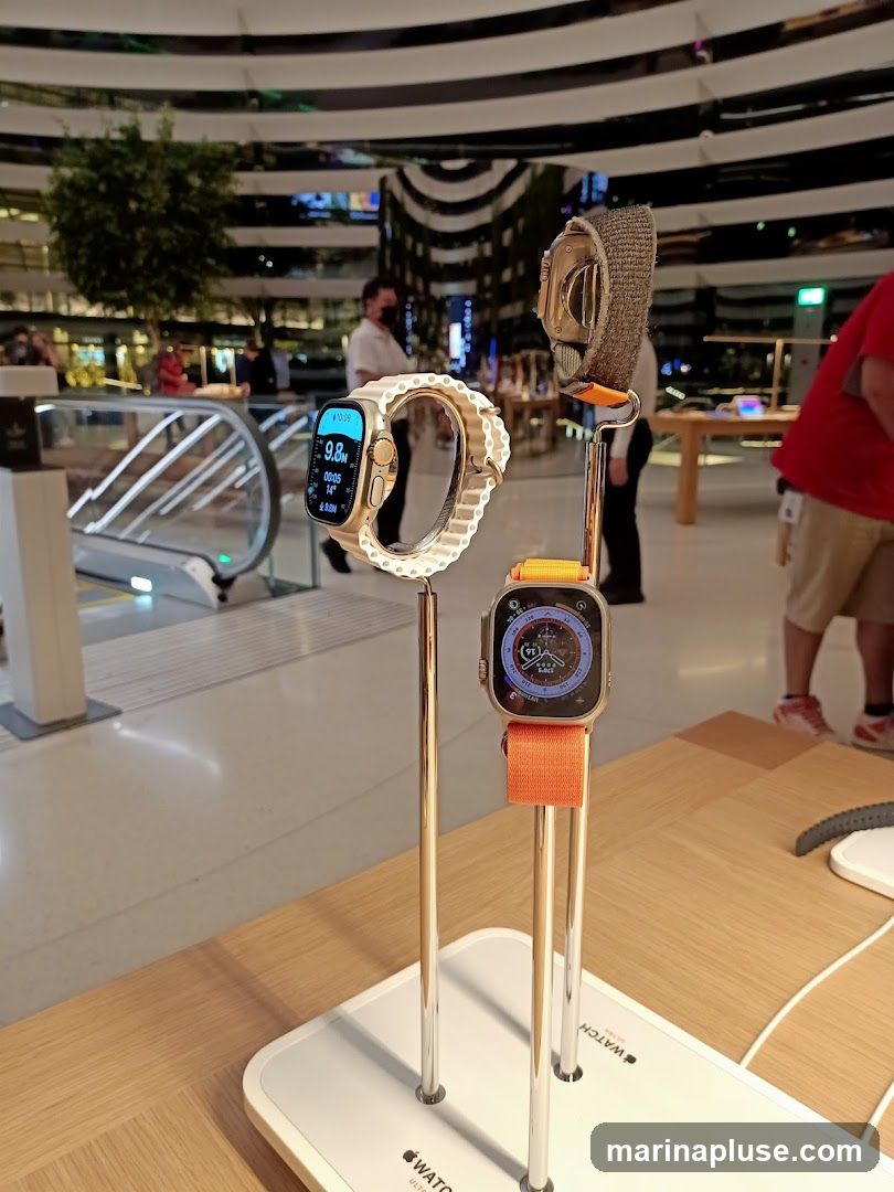 Two Apple Watches are displayed on stands in what appears to be a retail store. The watch on the left has a white band and displays time, date, and activity metrics. The watch on the right has an orange band and shows a compass and other data. In the background, escalators and shoppers are visible, suggesting a busy commercial environment. The Apple logo is visible on the display stands.