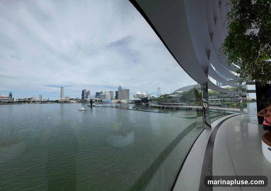 A panoramic view of a city skyline across a body of water, seen from inside a modern building with curved architecture and large windows. The water reflects the cloudy sky and the buildings. In the foreground, a woman is partially visible, seated on a bench.
