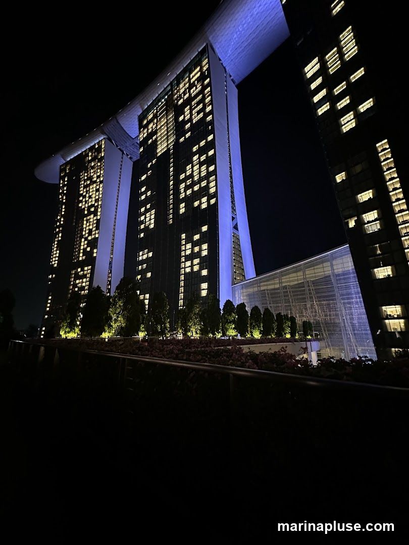 A nighttime view of the Marina Bay Sands hotel in Singapore, with its iconic three towers illuminated by lights. The sky is dark, and the building's windows glow with warm light. In the foreground, there are trees and landscaping, also lit up.