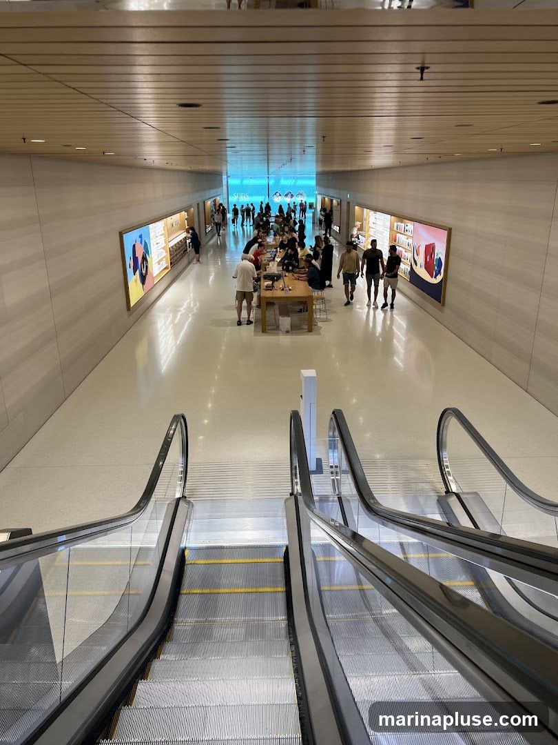 An overhead view of a brightly lit, modern retail store interior. Escalators lead down into the main shopping area, which features long display tables with customers interacting with products. Large, colorful advertisements are displayed on the walls. The ceiling is paneled with wood, and the floor is a polished white surface. The overall ambiance is clean, spacious, and contemporary.