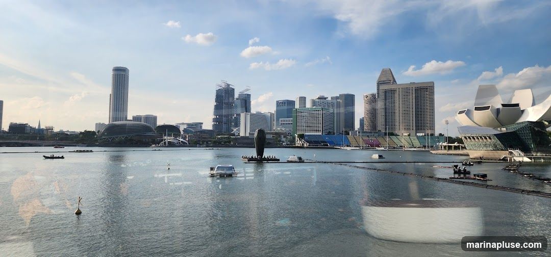 A panoramic view of the Singapore skyline, featuring modern skyscrapers and iconic landmarks such as the Marina Bay Sands resort and the ArtScience Museum. The image captures the bustling activity on the water, with boats and floating structures visible. The sky is partly cloudy, indicating a bright day.