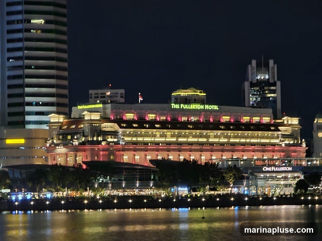A nighttime view of The Fullerton Hotel, a grand building illuminated with pink and yellow lights. The hotel's name is prominently displayed in green lettering on its facade. In the foreground, a body of water reflects the lights of the hotel and surrounding buildings. To the right, a sign reads "ONEFULLERTON".
